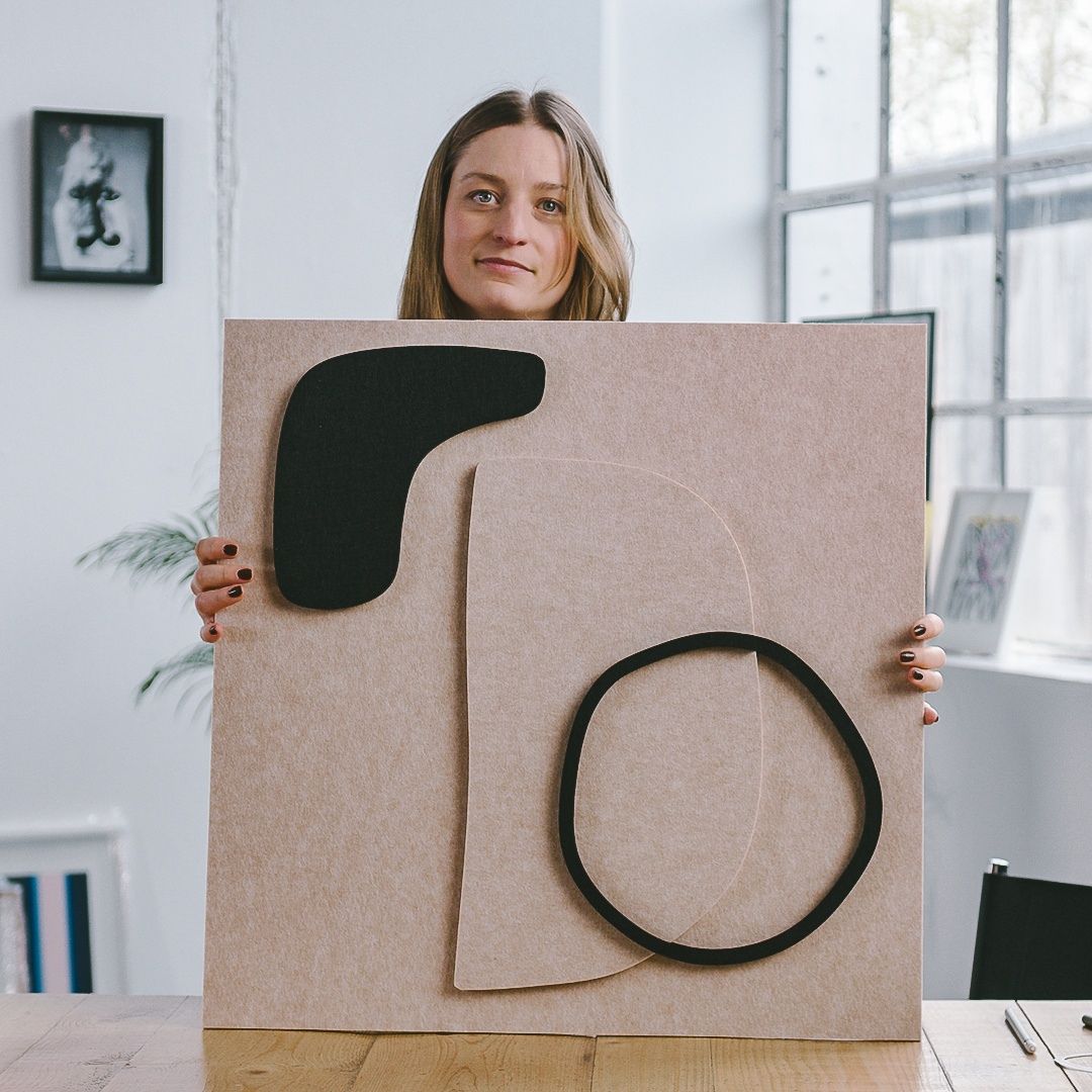 Designer Saskia Hübner holding a Nordic Bauhaus-inspired acoustic panel with minimalist black and beige geometric shapes.